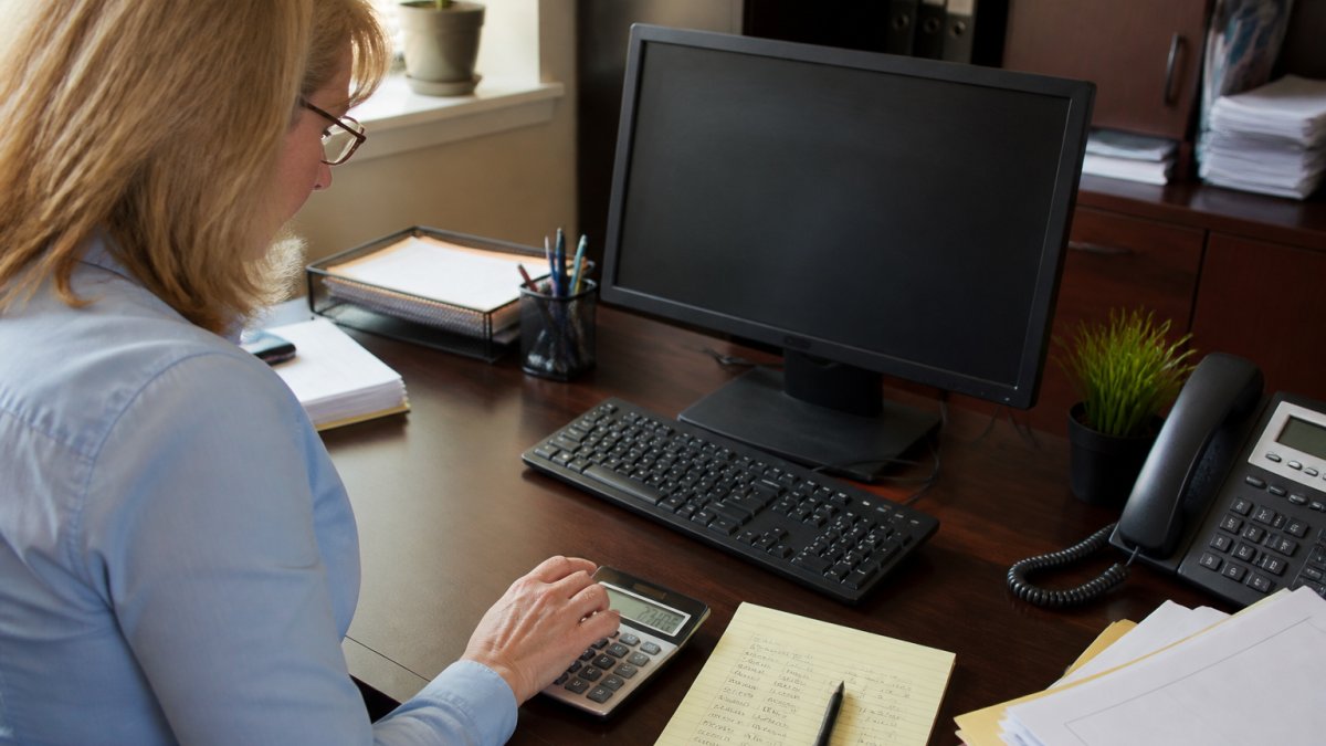Practice manager reviewing billing administrative costs on a monitor with handwritten notes and a calculator